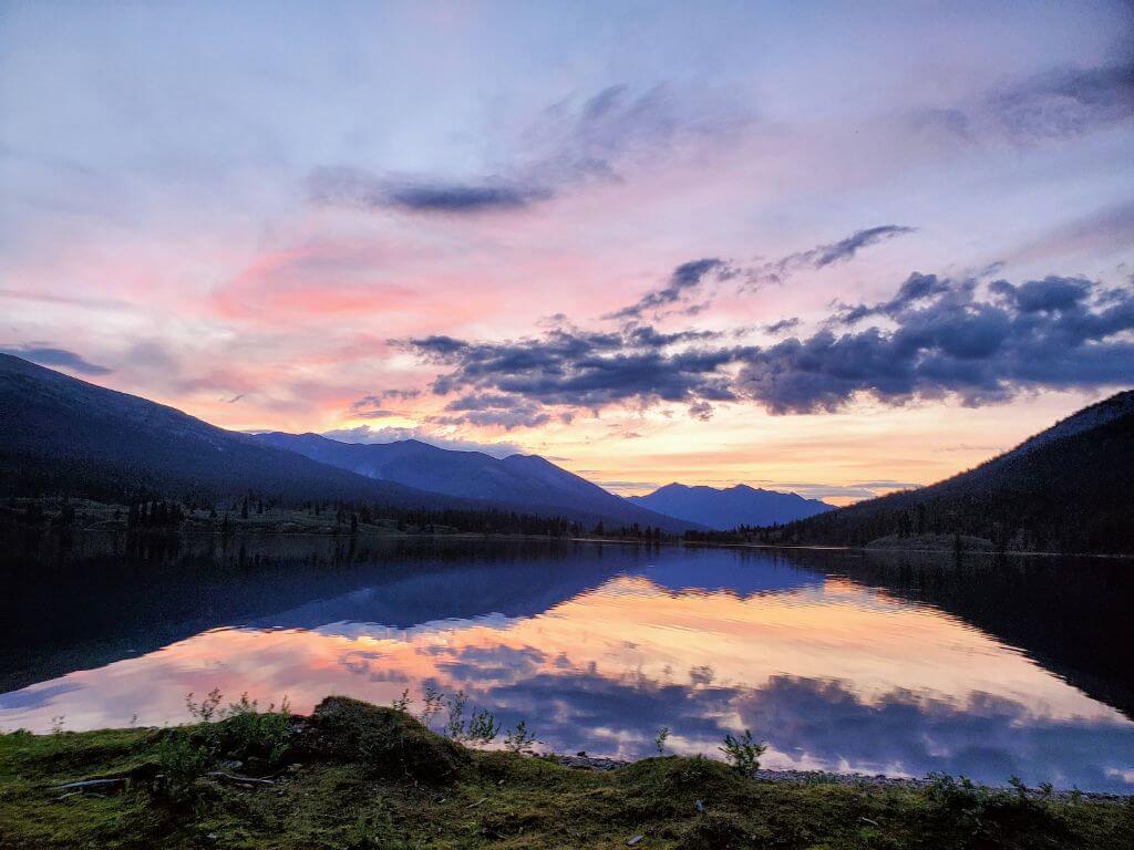 Sonnenuntergang am See im Yukon Territory in Kanada mit im See spiegelnden Bergen und Wolken.