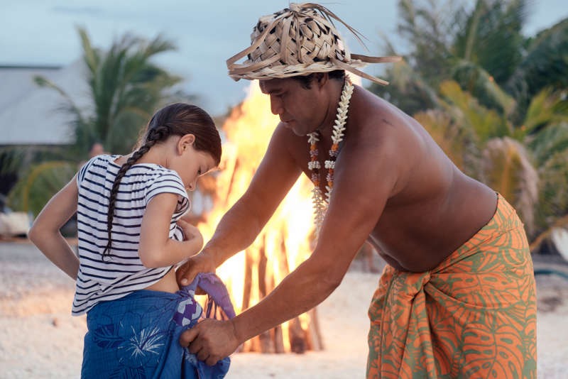 Polynesier zeigt Kind wie man einen Wickelrock wickelt. Lagerfeuer am Strand in Polynesien im Hintergrund